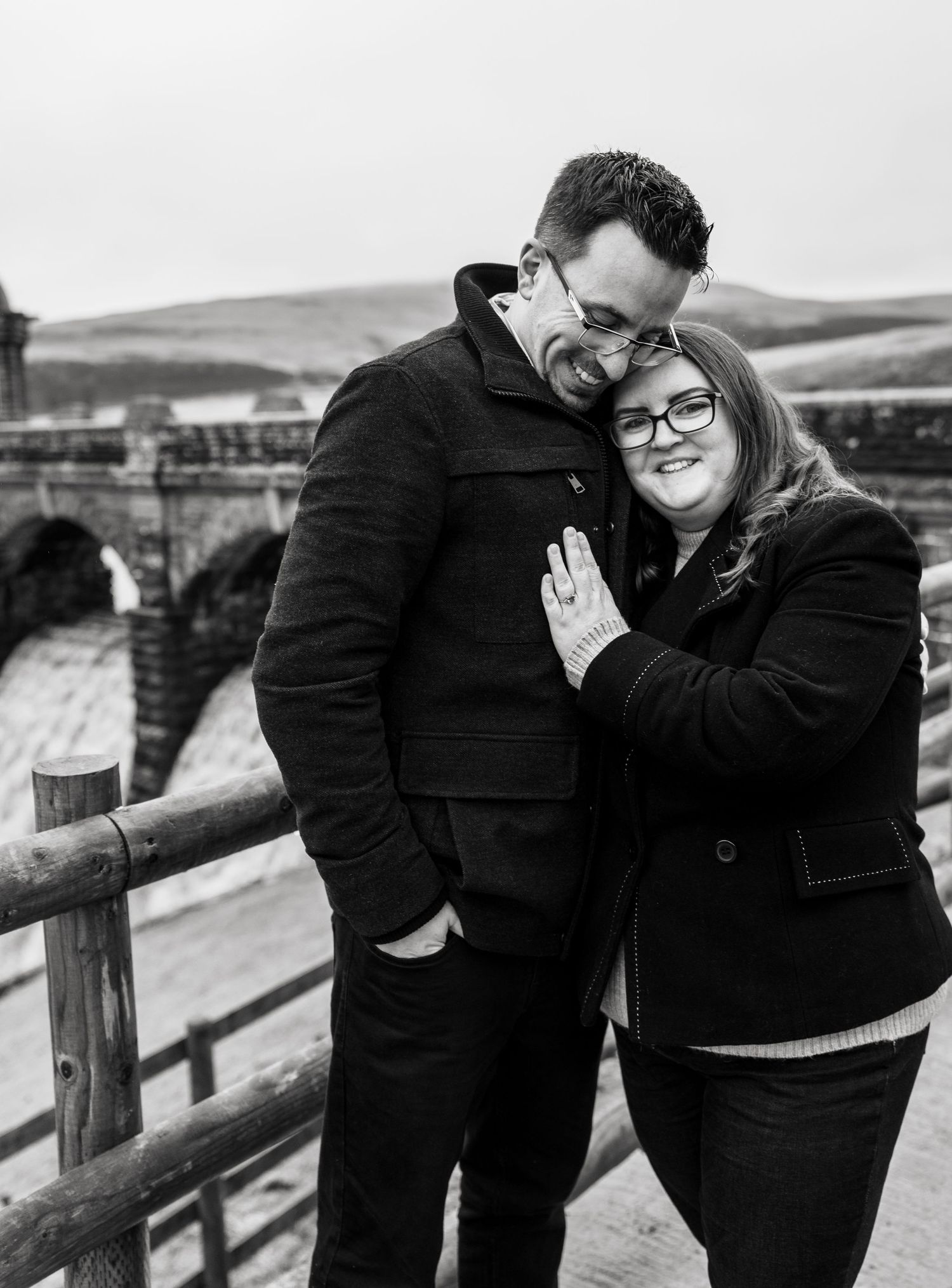 A couple in black winter coats share playful candid moments along a wooden railing during an outdoor photoshoot.