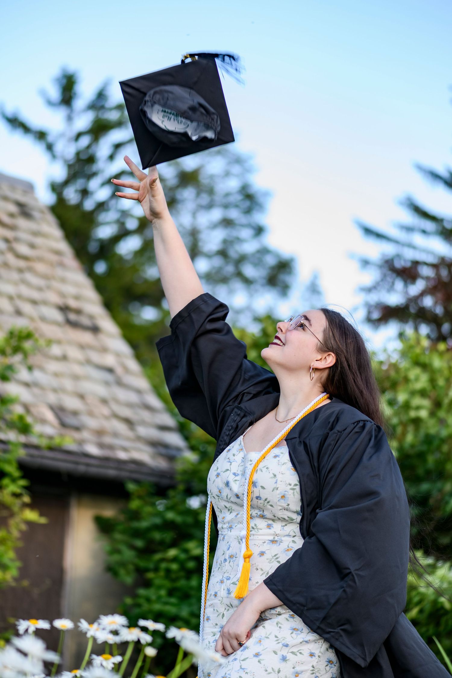 A female graduate tossing her cap up in the air and smiling near a cottage and flowers
