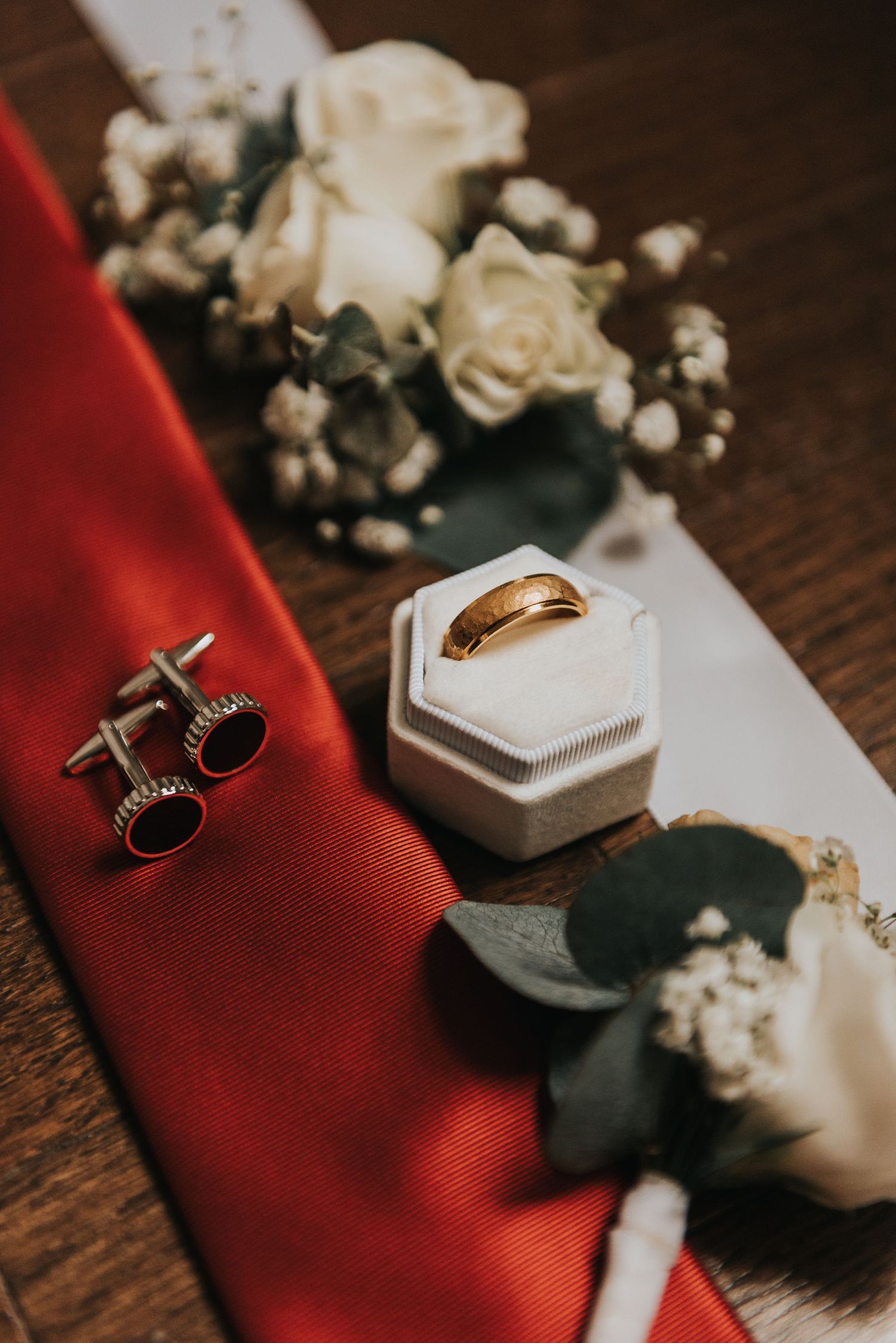Close-up of wedding rings, red tie, and white floral boutonniere displayed on wooden surface with hexagonal box.
