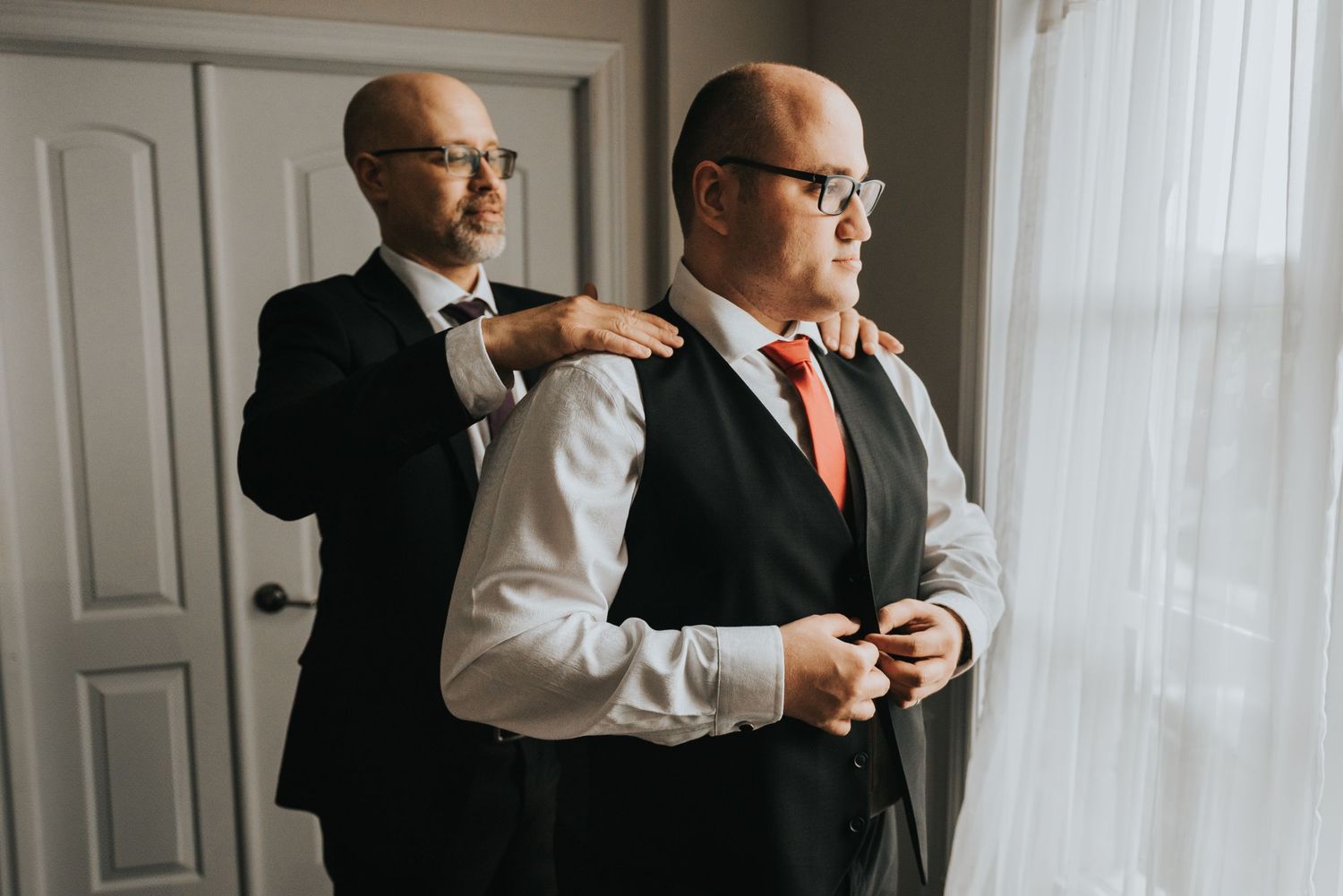 A groom in a black suit and glasses checks his phone while getting ready in a bright room with white walls.