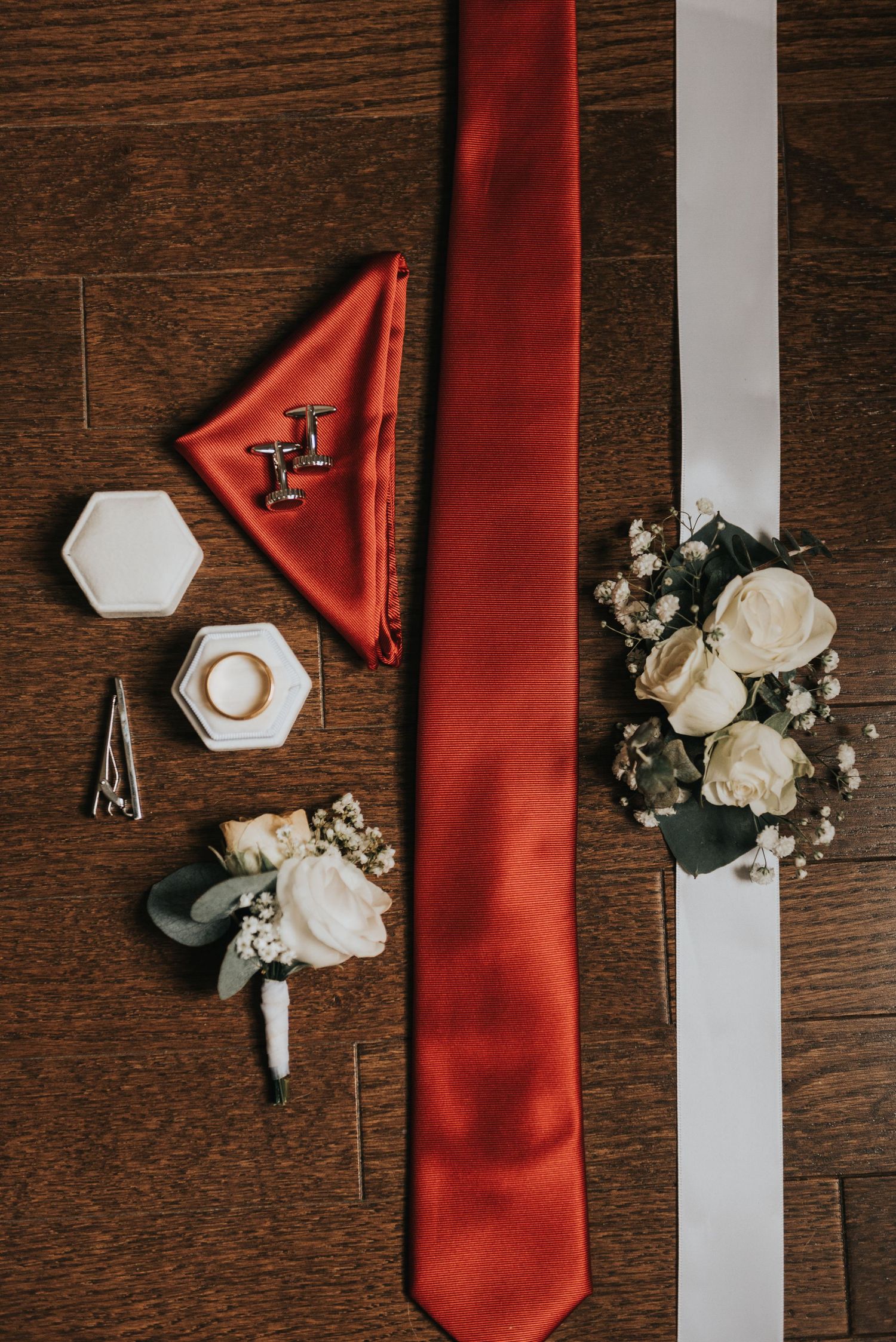 Wedding accessories including a red tie, white boutonniere, and hexagonal ring box arranged on dark wooden surface.