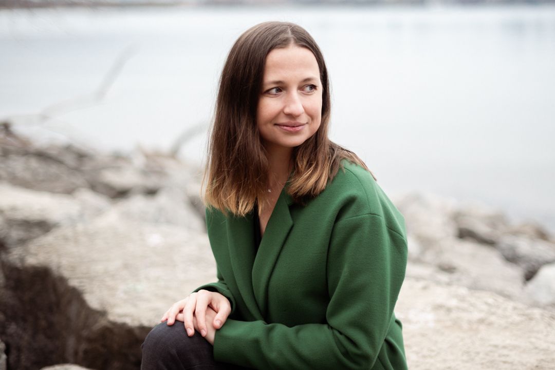 Person wearing green coat sits thoughtfully by rocky waterfront.