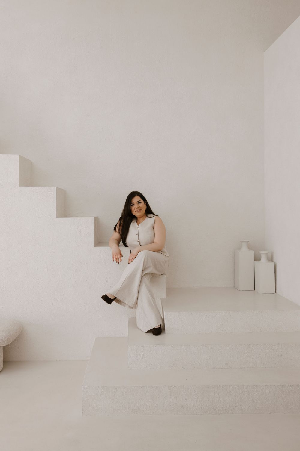 Minimalist white interior featuring stairs and decorative vases with person seated in neutral-toned outfit.