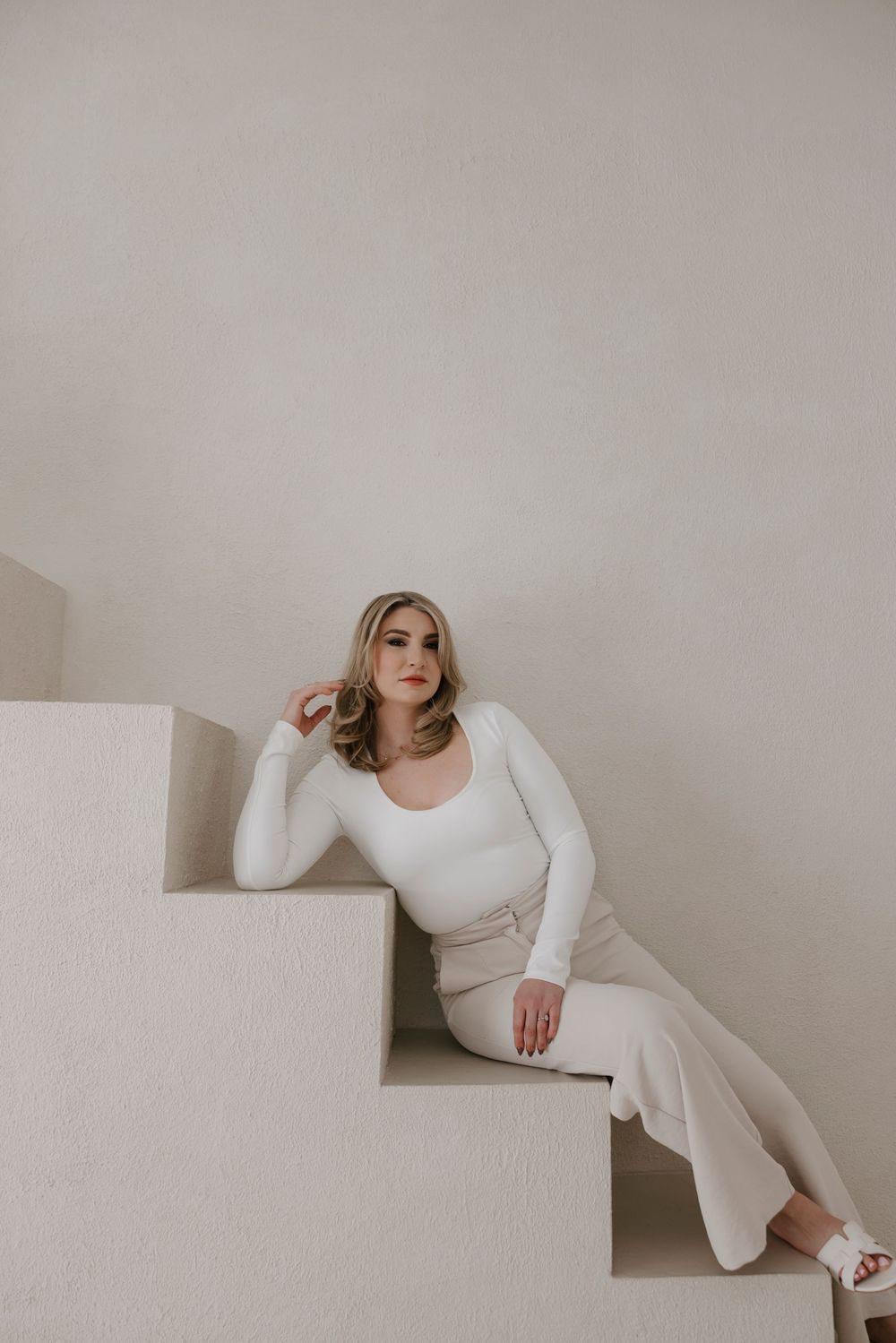 Model in cream colored outfit reclines on white architectural steps in minimalist setting.