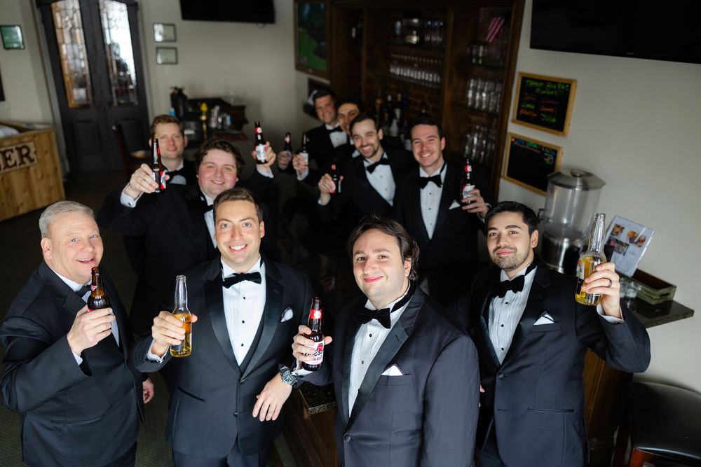 A group of groomsmen in black tuxedos and bow ties raise their drinks for a celebratory toast at a bar.