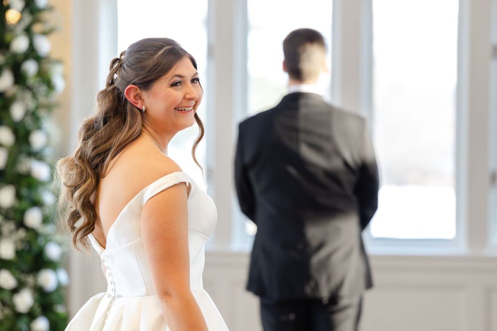 A bride in a white sleeveless wedding dress smiles while standing in an elegant indoor venue with floral decorations.