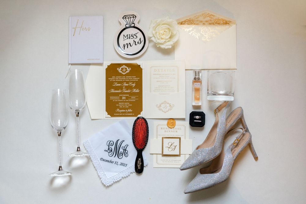 Elegant wedding accessories including invitations, rings, shoes, and perfume arranged on white surface in soft lighting.