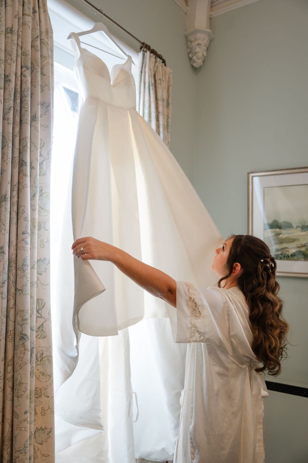 A white wedding dress hangs by a window while someone in a silk robe reaches up to touch it.