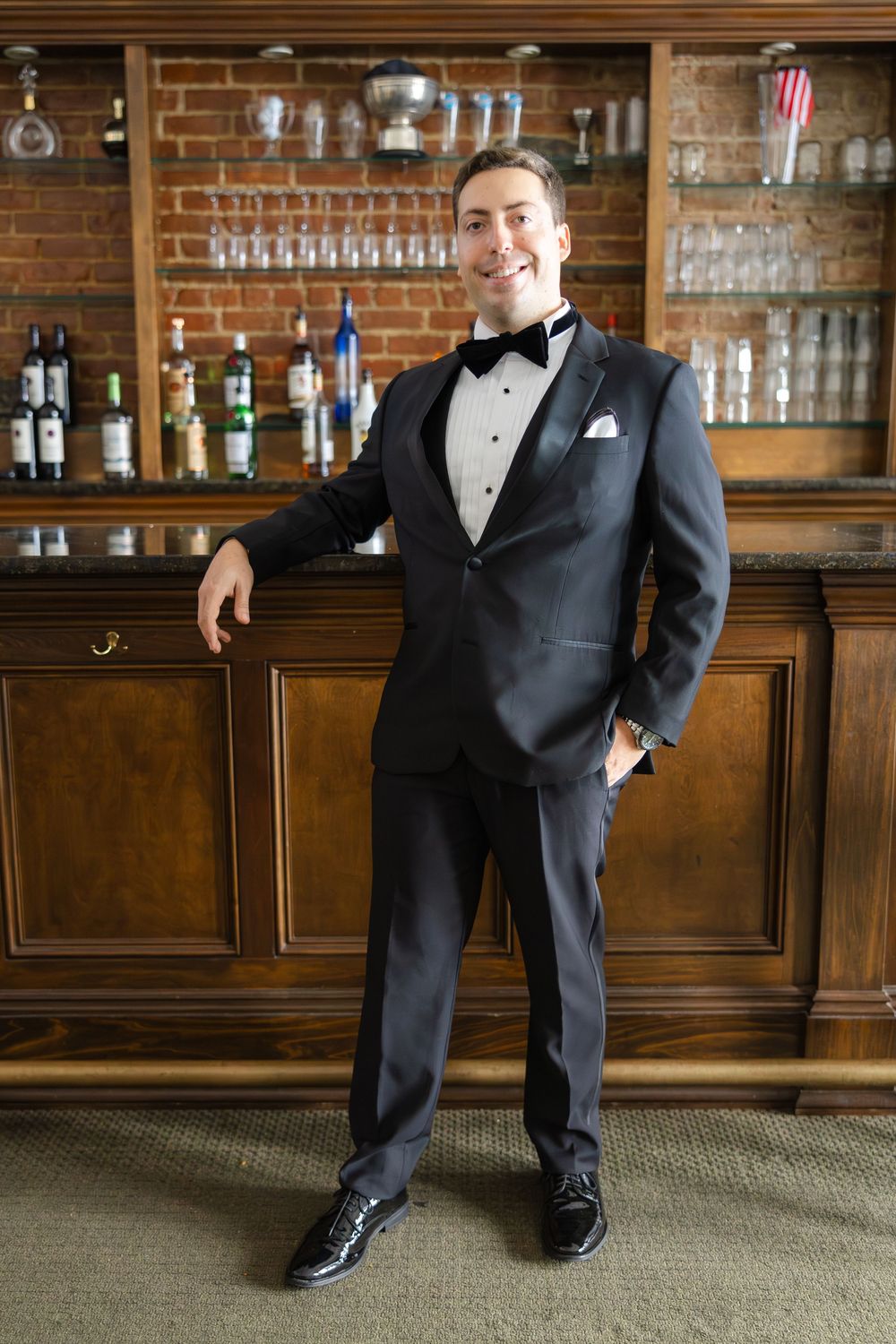 A person in a black tuxedo and bow tie poses casually against a wooden bar with glassware displayed in the background.