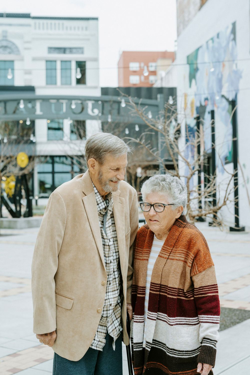 Two people walking together and sharing a warm moment on a city sidewalk with buildings in the background.