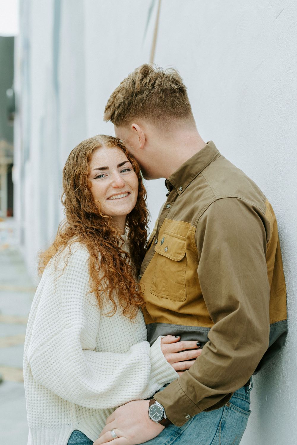 A couple shares an intimate moment against a white wall, with her wearing a cozy white sweater and him in a brown jacket.