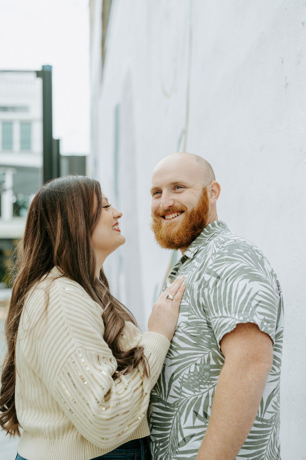 A couple in matching palm print shirts share playful laughter and affection against a white building exterior.