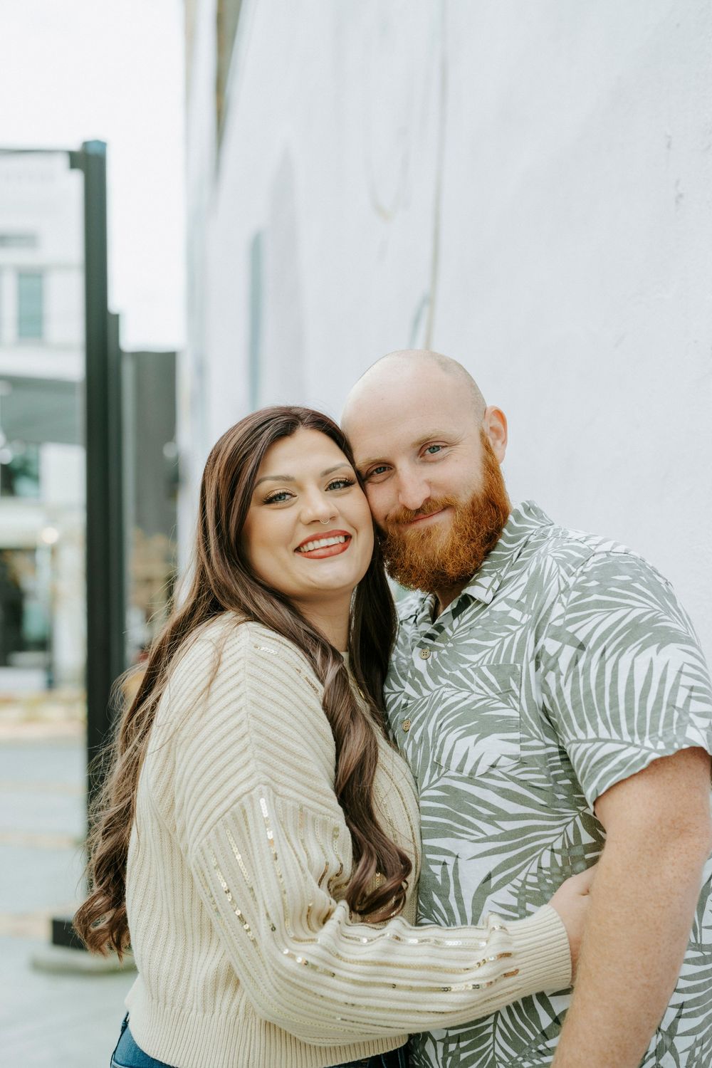 A couple in coordinating outfits smile together on a city street, with one wearing a cream sweater and the other a palm print shirt.