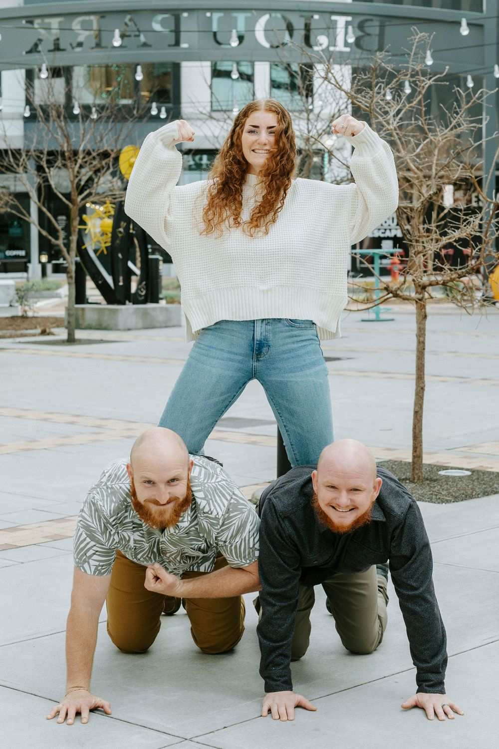Playful outdoor photo with two people doing push-ups while another stands on their backs in a white sweater.