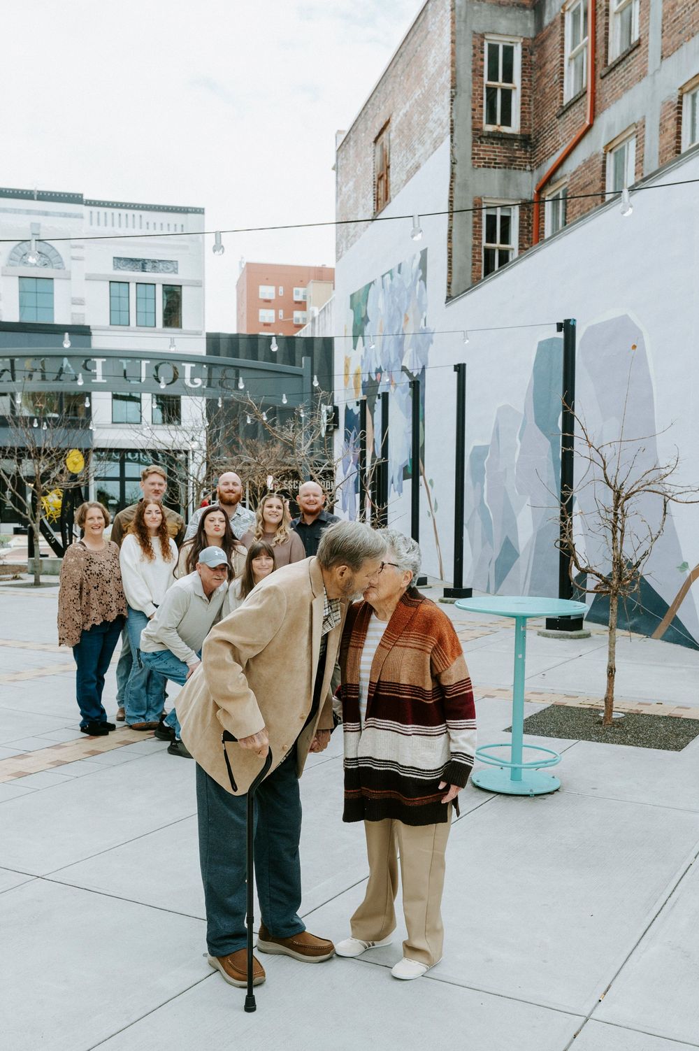 A group of people in winter coats stand together outdoors in front of a brick building on a cloudy day.