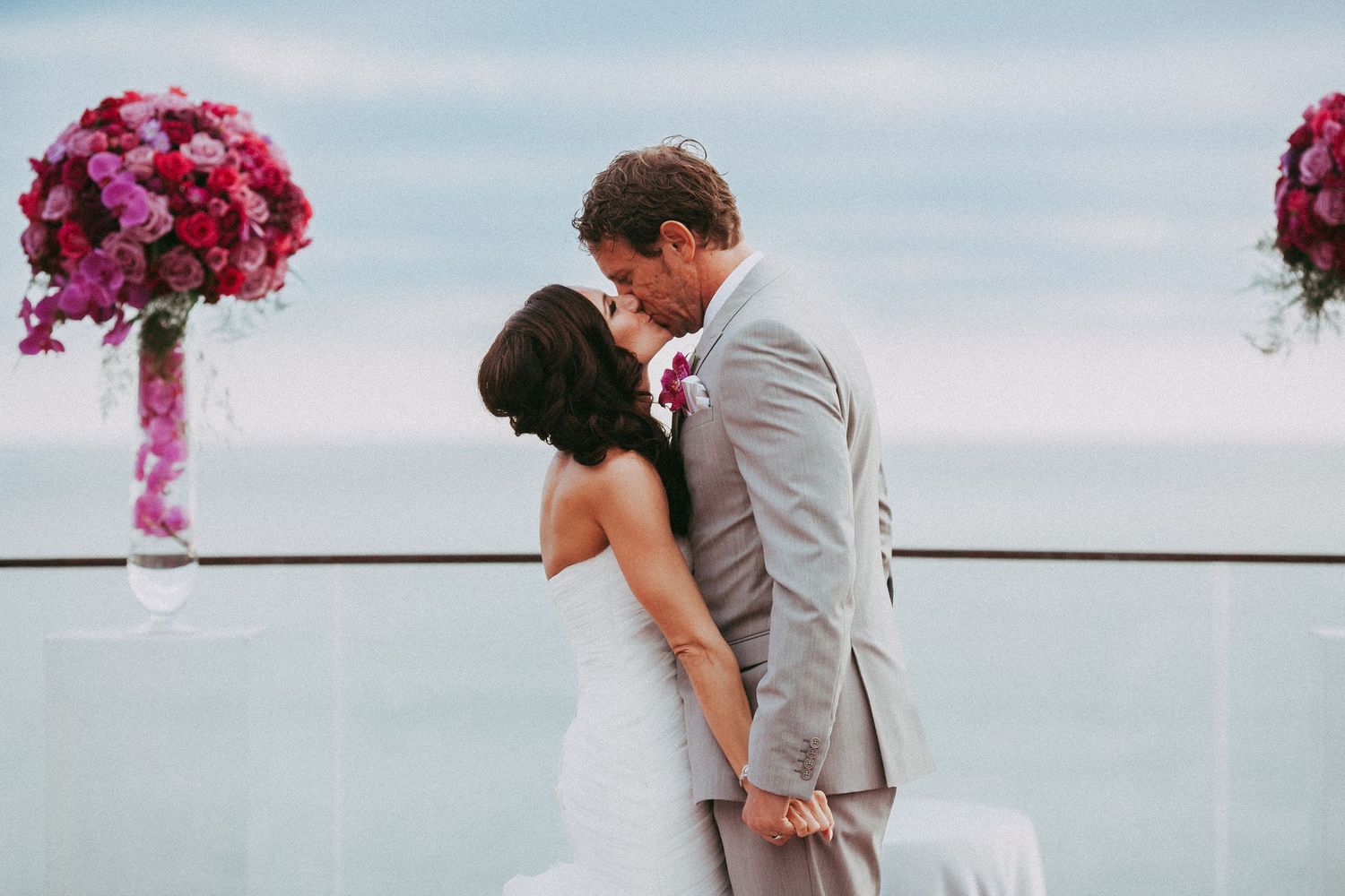 Couple embraces in romantic kiss against ocean backdrop with vibrant pink floral decorations.
