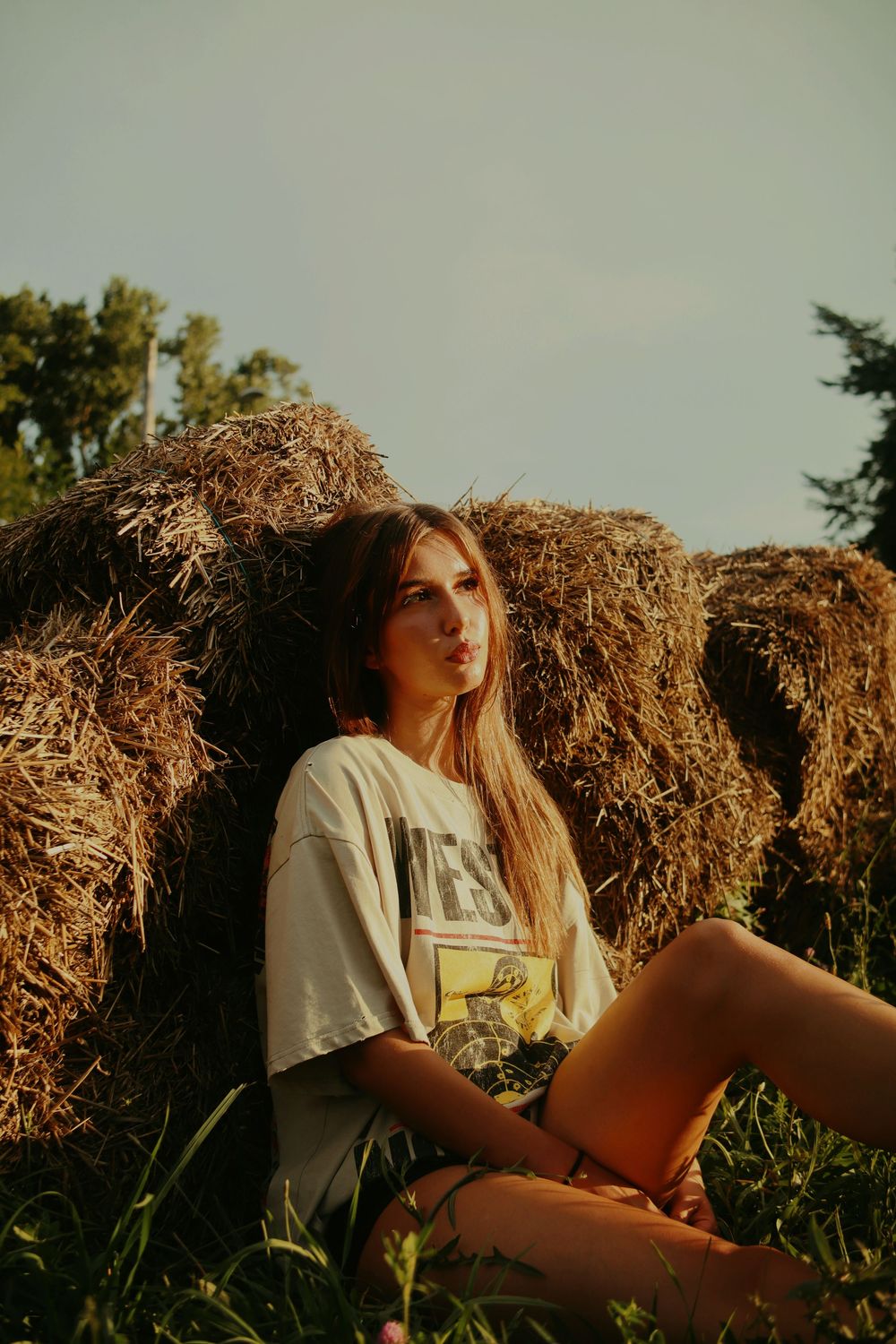 A person in a white t-shirt sits near hay bales during golden hour in a rustic countryside setting.