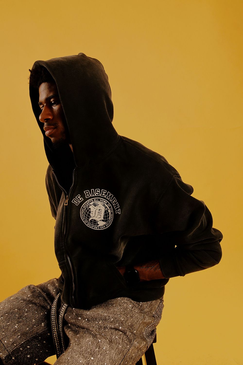 Figure in black hooded sweatshirt with logo poses against golden yellow background in dramatic lighting.