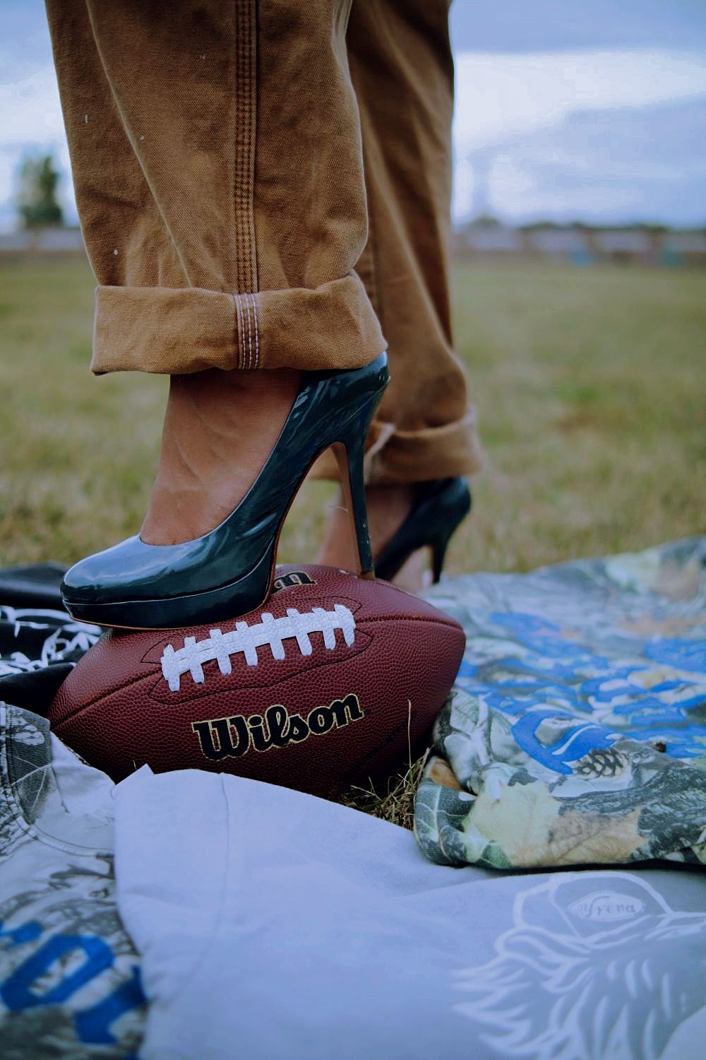 A foot wearing a black shoe steps on a Wilson football resting on a patterned blanket outdoors.