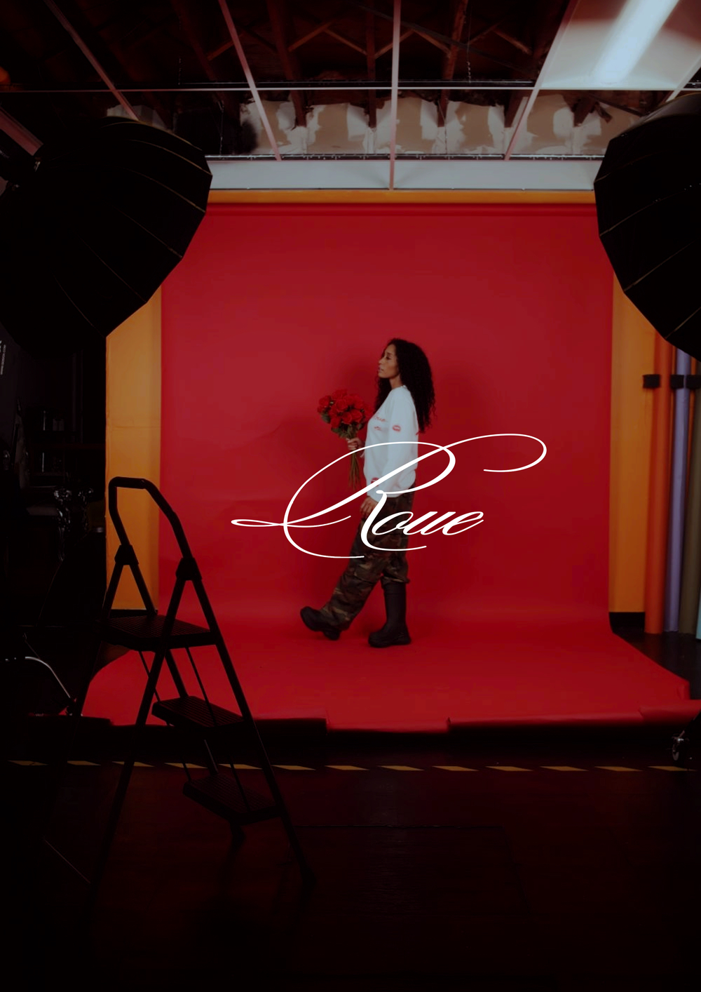 A person poses against a vibrant red backdrop in a photo studio with lighting equipment and a stepladder visible.