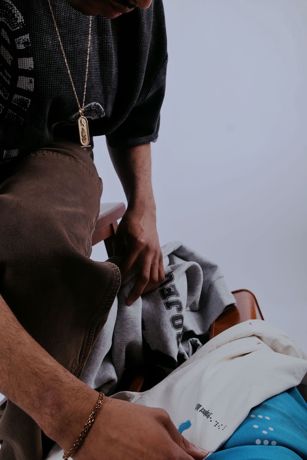 Man wearing gold chain necklace and bracelet folding gray and white clothing on a light background.