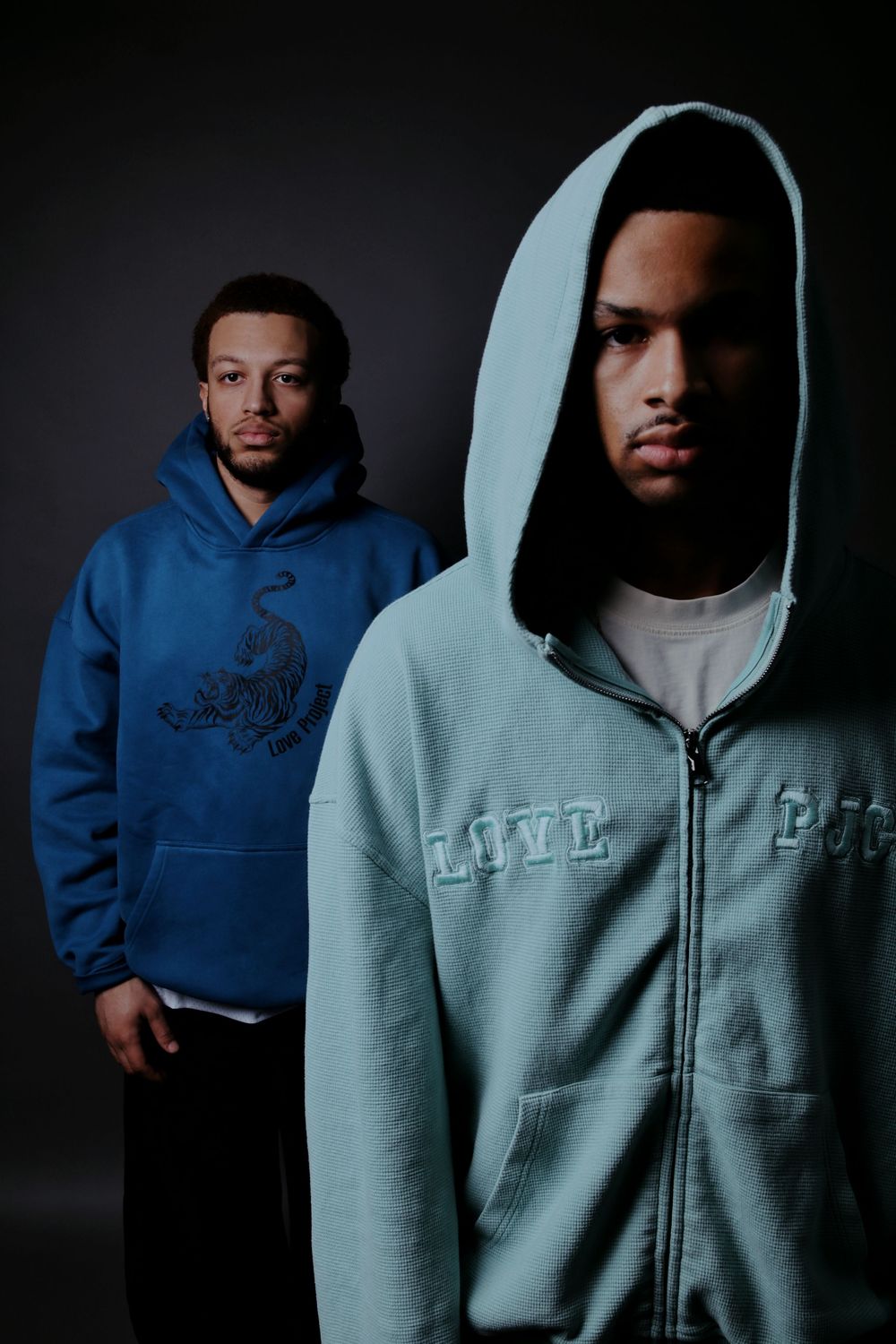 Two young men in hoodies pose against dark background, one in blue, one in light blue zip-up hoodie.