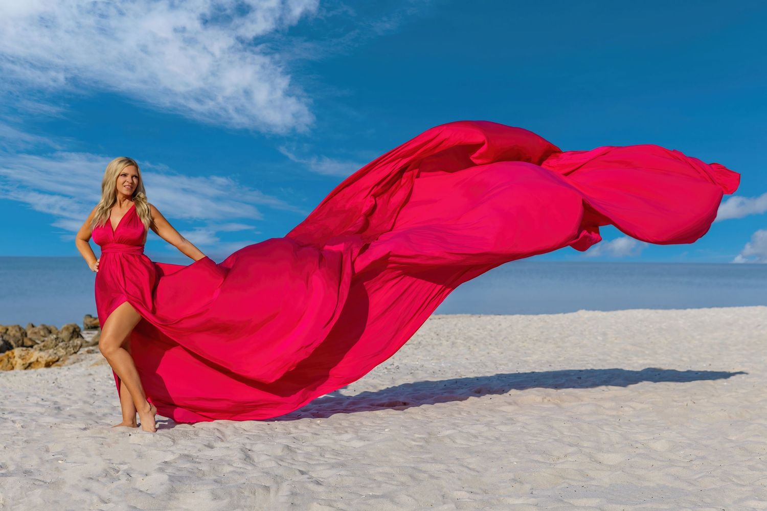 A flowing red dress billows dramatically against white sand dunes and blue sky in a desert landscape.