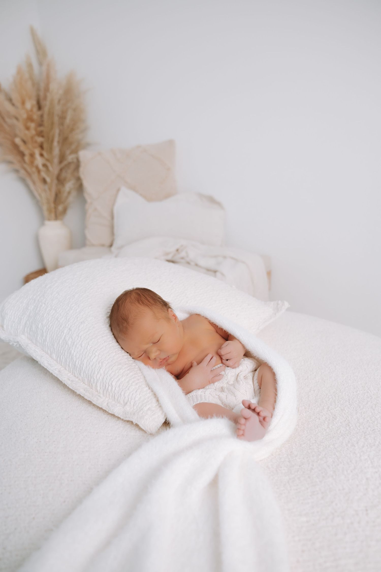 A sleeping newborn baby wrapped in soft white blankets on a white bed with dried wheat decor in the background.