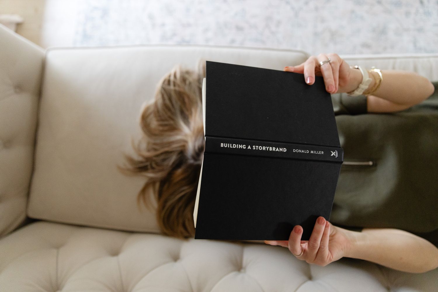Laura Pulliam lying on a beige couch reading a black hardcover book while relaxing at Makai Creative Studio in Spring Texas.