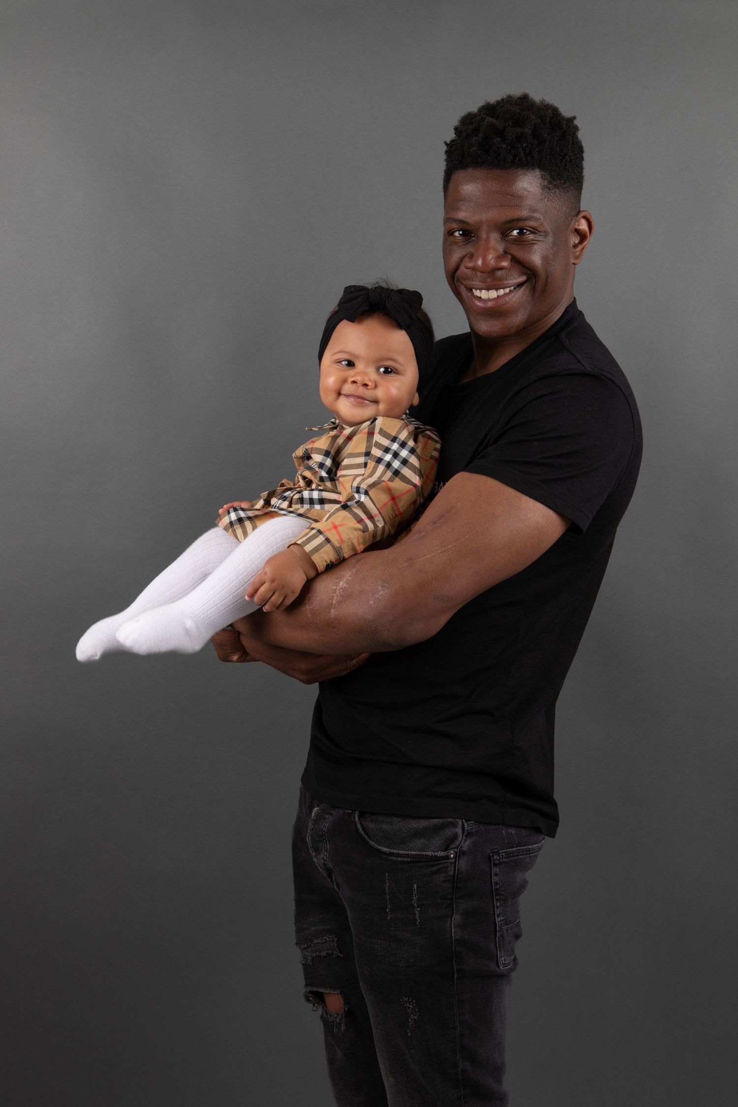 A parent in black clothing holds a smiling baby wearing a plaid shirt and white pants against a grey studio backdrop.