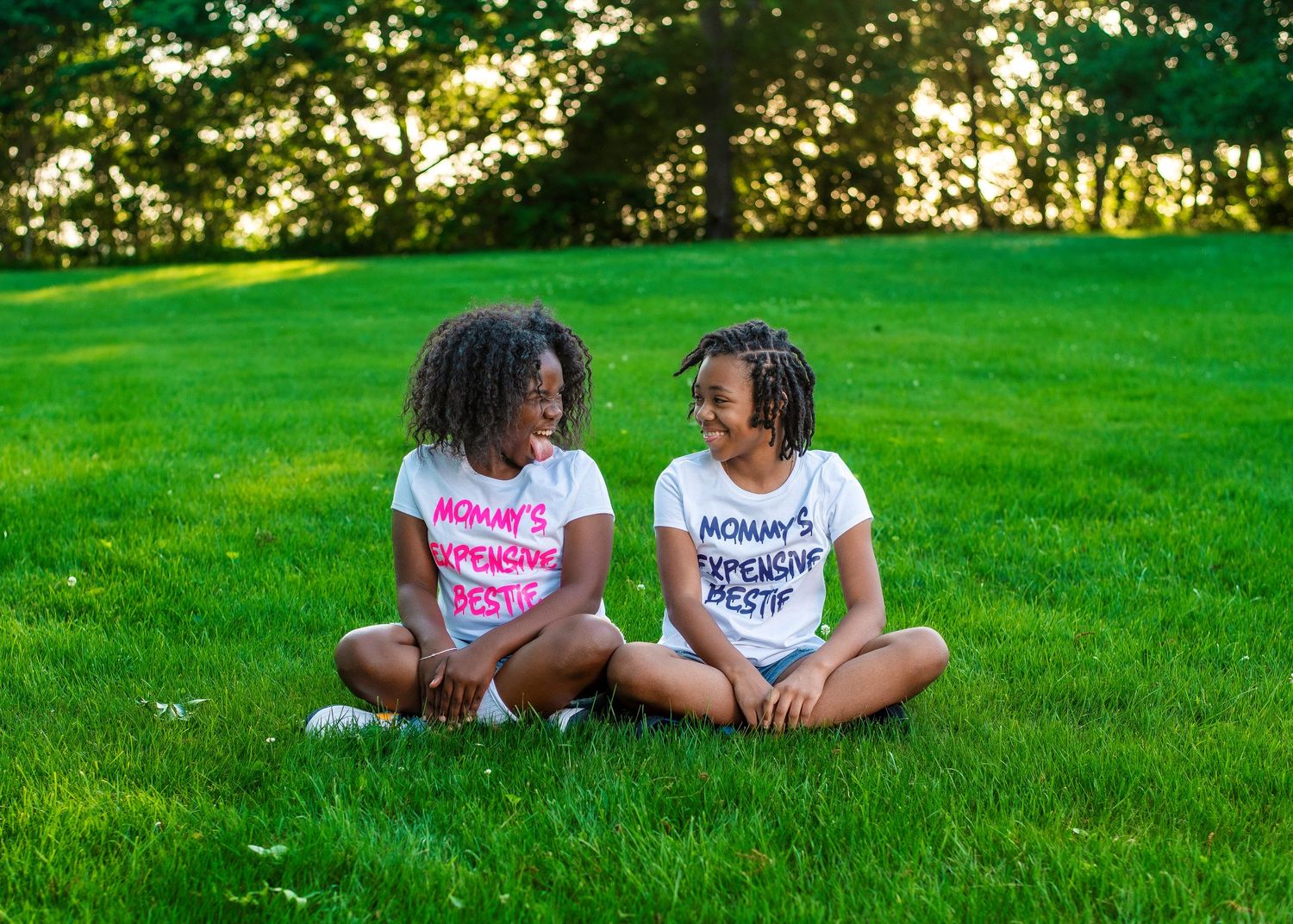 Two people sit together on lush green grass wearing matching white t-shirts with pink and blue text.