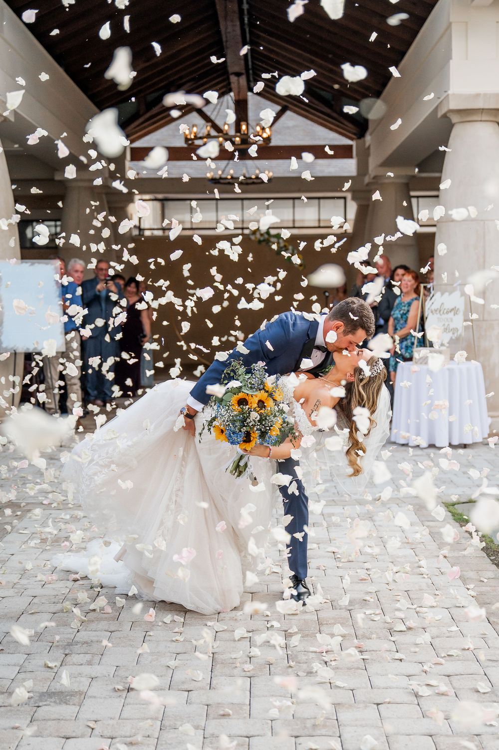 White flower petals scatter through the air during a joyful wedding celebration on a stone patio.