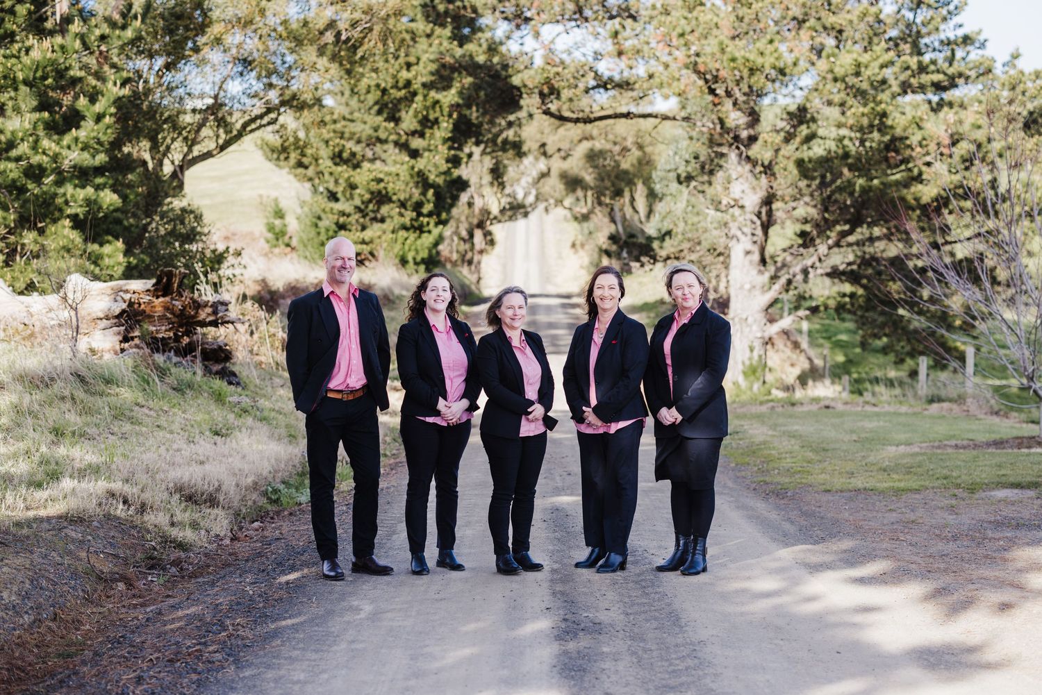 Wedding party in dark formal attire poses together on rustic outdoor path in natural setting.