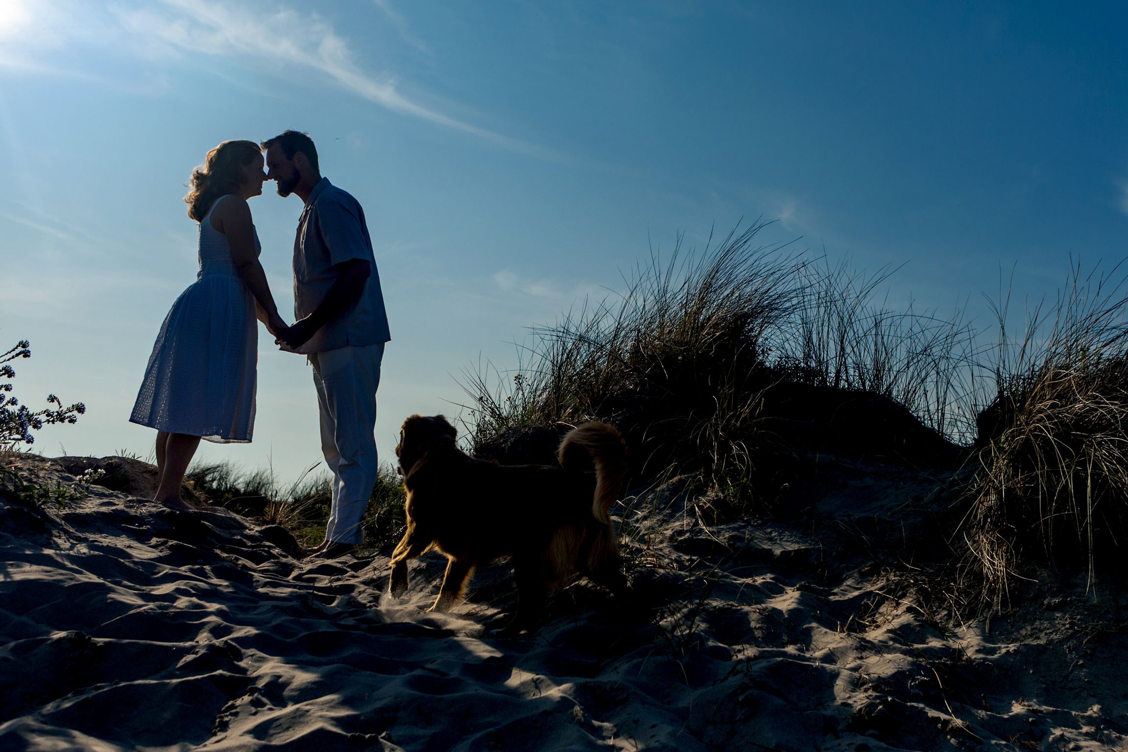 Koppel in silhouet op een duin bij Wijk aan Zee met hun hond spelend op de achtergrond, vastgelegd door Luzan Veenendaal Fotografie