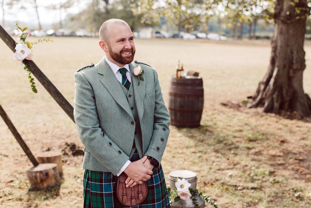 A couple in green jackets and kilts holds hands during an outdoor wedding ceremony in a rustic setting.