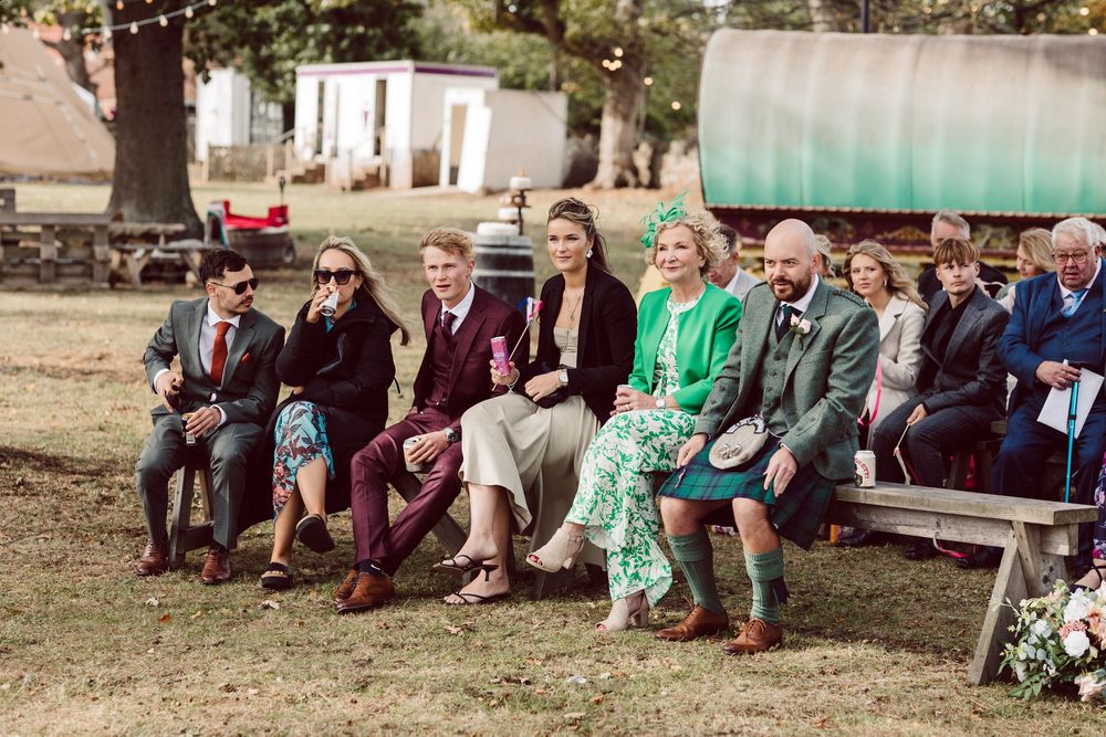 Wedding guests sit on wooden benches in a rustic outdoor setting for a ceremony.