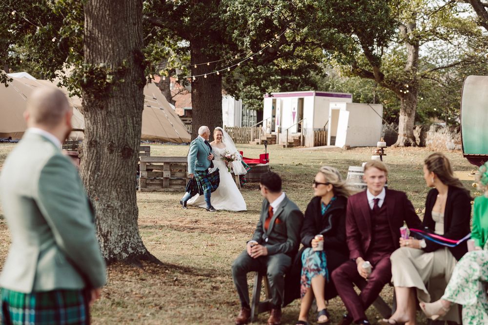 Wedding guests gather outdoors under large trees during a rustic countryside celebration in formal attire.