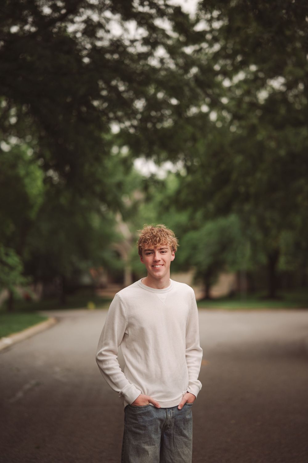 Smiling teenage boy with curly blonde hair wearing a white sweater and jeans standing on a tree-lined street.