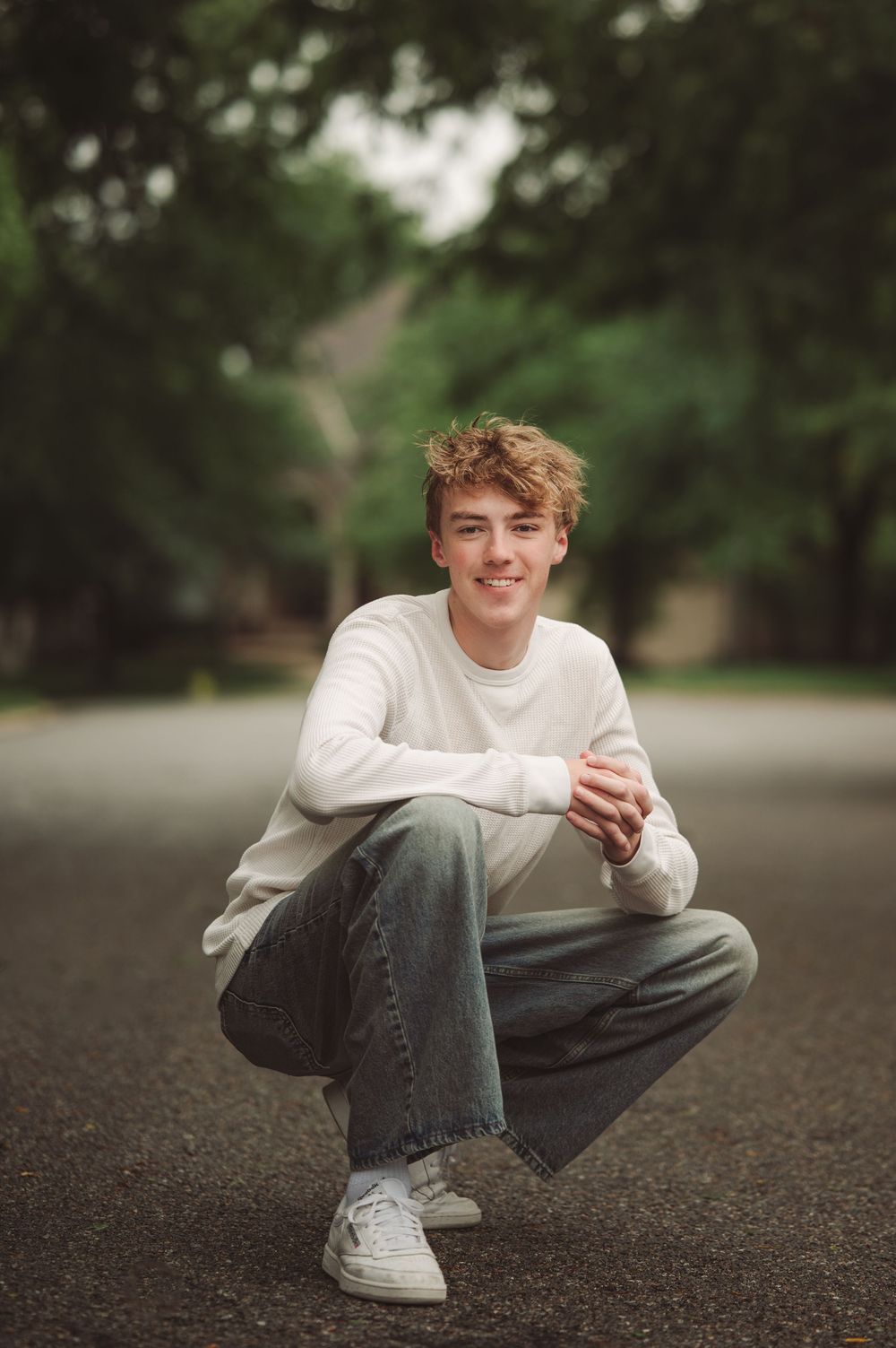 Teenage boy with curly blonde hair crouching on a road wearing a white sweater and jeans with white sneakers.