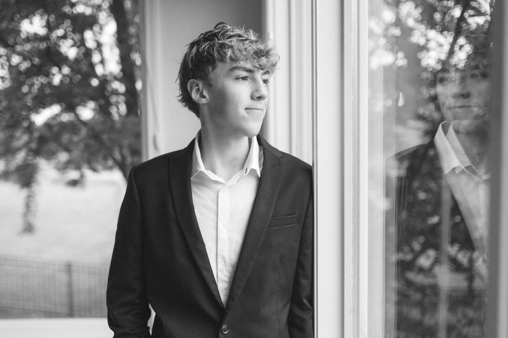 Black and white portrait of a teenage boy in a blazer gazing out a window with his reflection visible.
