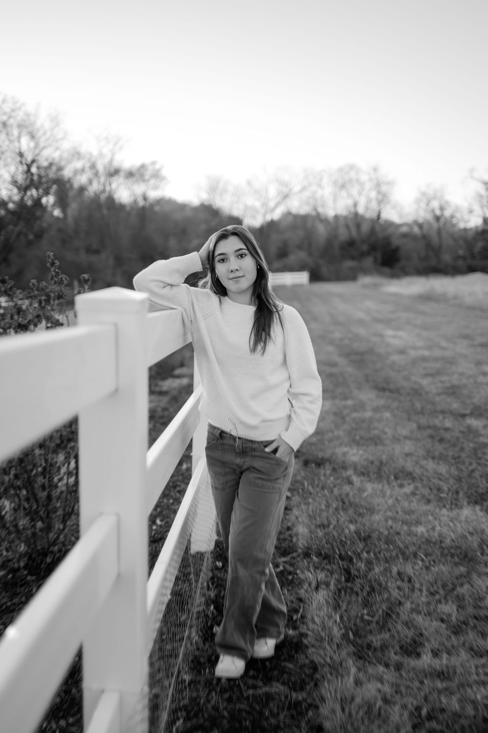 Black and white senior portrait of teenage girl leaning against white fence in open field.