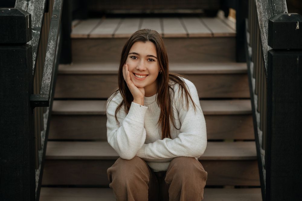 Smiling teenage girl in white sweater sitting on wooden steps with chin resting on hands.