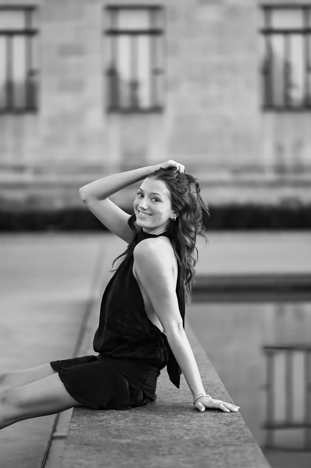 Black and white photo of a smiling young woman in a black dress posing on a ledge outdoors.