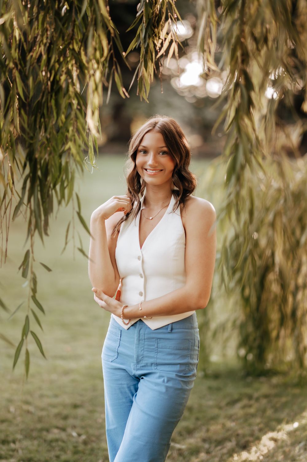 Smiling brunette woman in a white vest top and jeans posing under hanging willow tree branches.