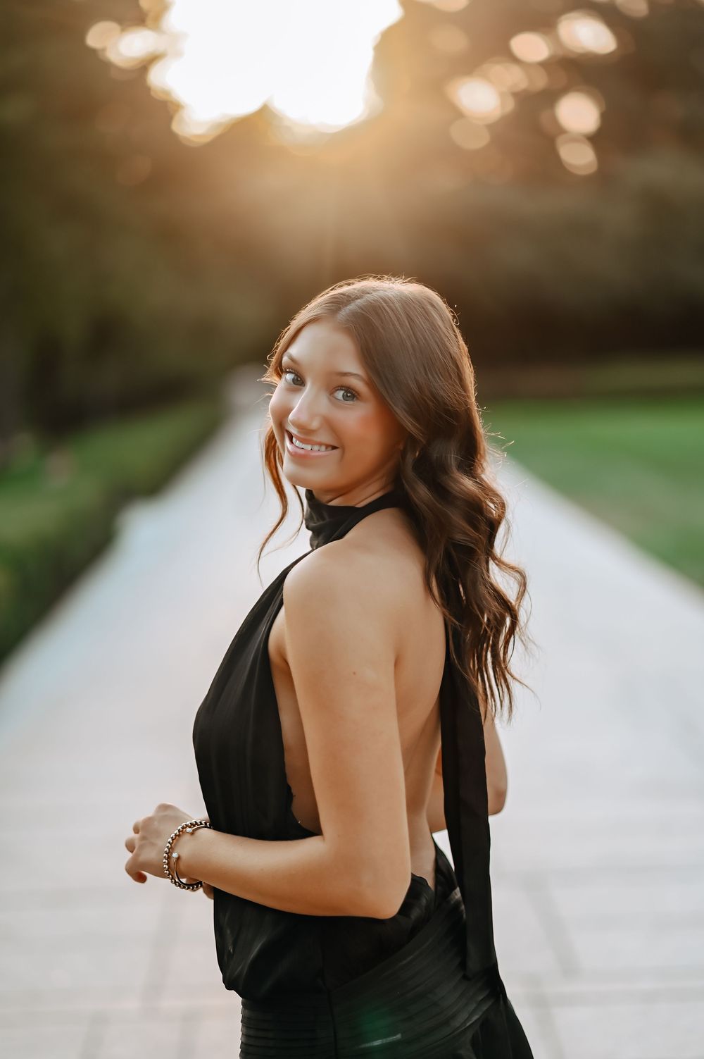 Young brunette woman in a backless black halter dress smiling over her shoulder at golden sunset.