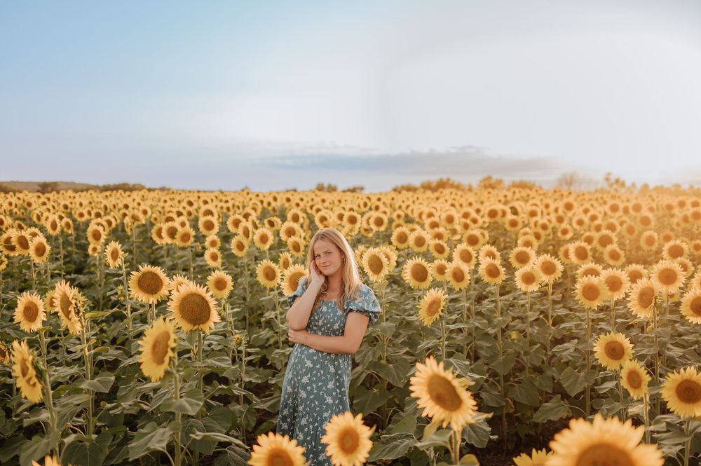 Blonde woman in a blue floral dress posing among rows of sunflowers in a wide open field.
