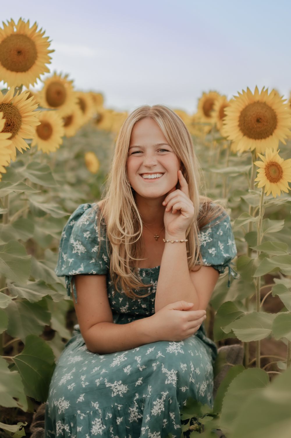 Smiling teenage girl with blonde hair sitting among sunflowers wearing a blue floral dress outdoors.