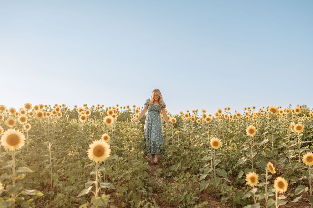 Young woman in a floral dress standing in a vast sunflower field under a clear blue sky.