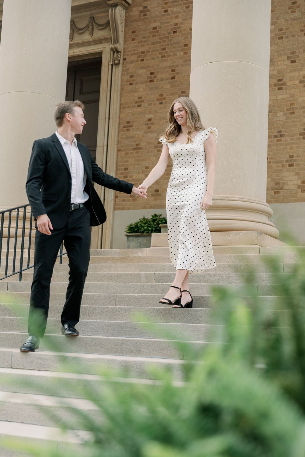 A couple in formal attire shares a playful moment on stone steps of a brick building.