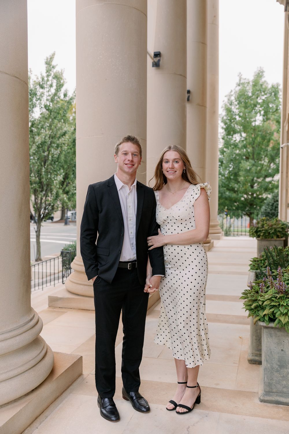 A couple poses together on a classic columned porch, with the person in a polka dot dress and another in formal attire.